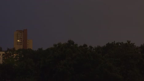 lightning bolt strike over urban city area surrounded by trees. flash of thunderbolt light on a rainy dark night. climate change.