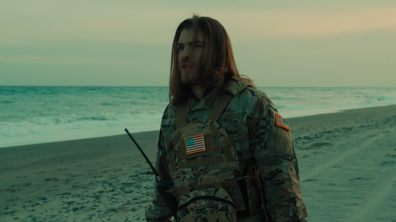 Militar charmoso com cabelo comprido andando na praia perto do oceano