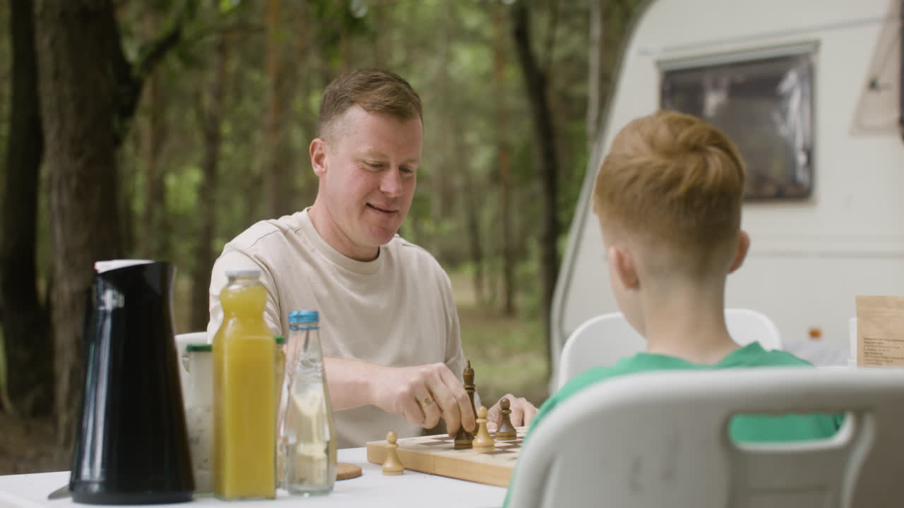 padre e hijo jugando al ajedrez sentados a la mesa en el campamento en el bosque