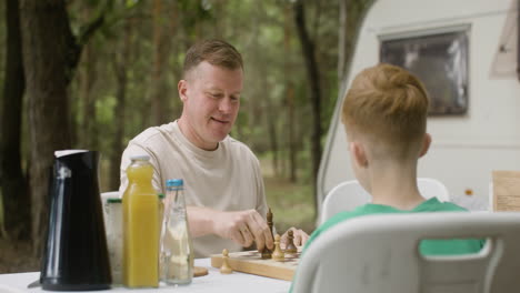 father and son playing chess while sitting at table at the camping in the forest