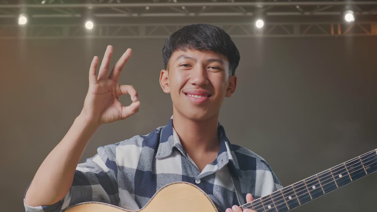 기타를 들고 있는 아시아 청소년의 클로즈업은 무대에서 은 몸짓을 하고 미소 짓는 모습을 보여준다.