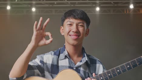 un primer plano de un adolescente asiático con una guitarra mostrando un gesto correcto y sonriendo en el escenario.
