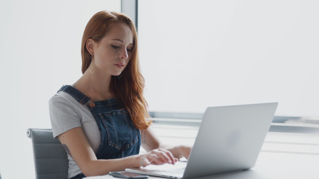 일상적으로 옷을 입은 젊은 사업가 여성이 현대적인 직장에서 책상에서 노트북에서 일하고 있습니다.