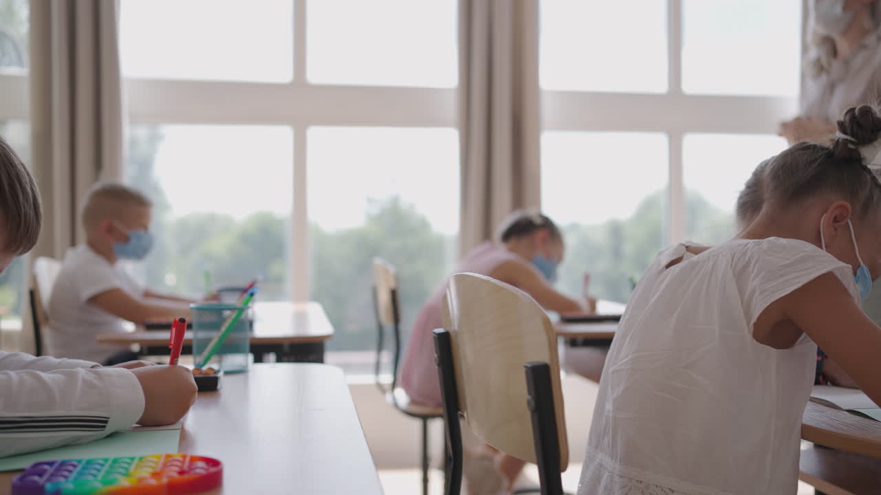 교실에서 선생님의 설명을 듣고 글을 쓰는 다민족 그룹의 아이들이 얼굴 마스크를 쓰고 수업에서 일하고 있습니다.