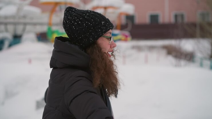 Athletic girl in black winter coat, knit hat, scarf, and glasses enjoying outdoor snow day, side profile view with curly hair and smiling face, expressing cheerful energy and positive winter lifestyle