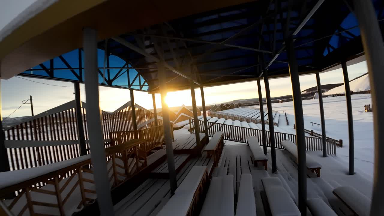 Ein kinoreifer Drohnenflug bewegt sich auf die glühende Sonne über schneebedeckten Tribünen zu. Warmes goldenes Licht kontrastiert mit der kalten Winterlandschaft und schafft eine friedliche und dramatische Luftaufnahme.