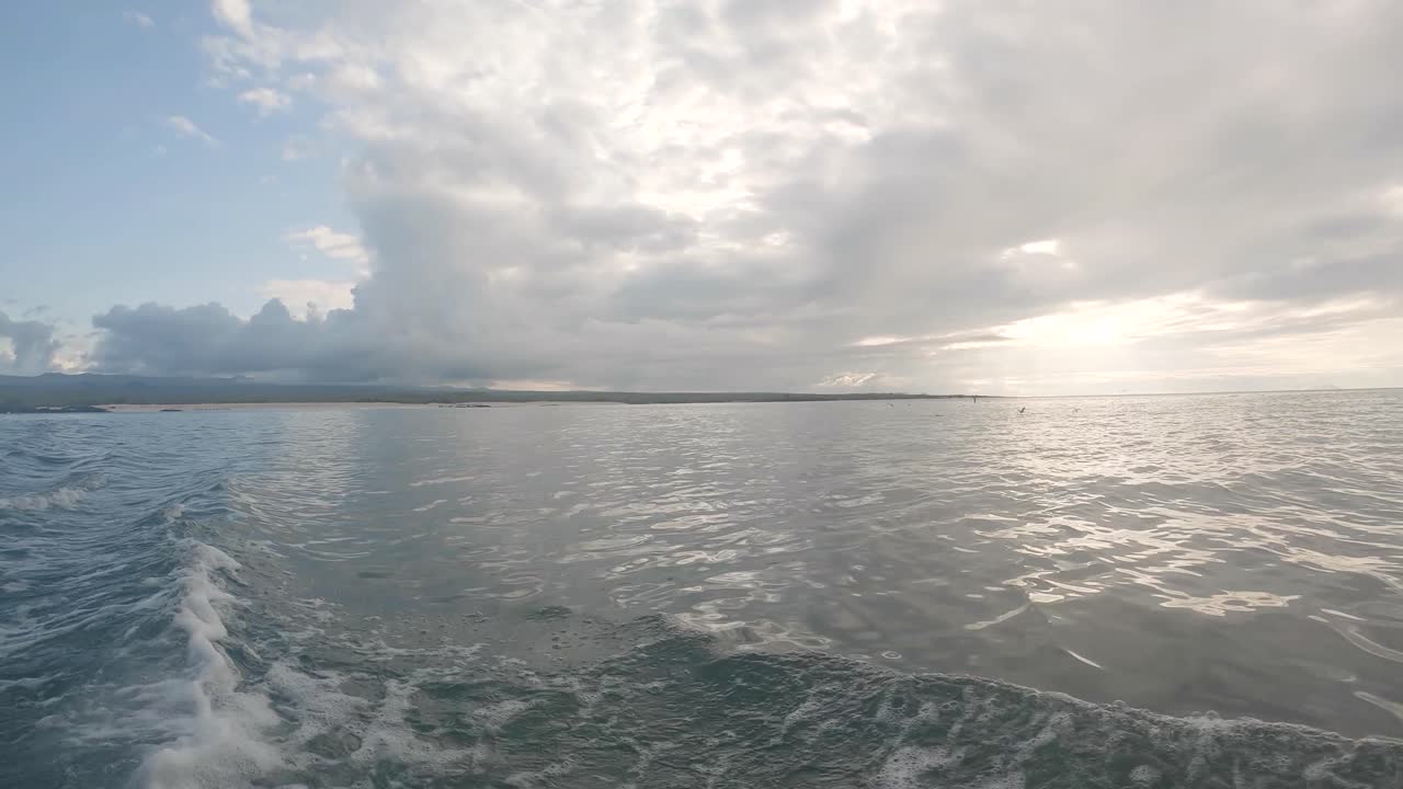 Some birds are flying close to the sea near an island of the Galapagos islands. There are waves created by the movement of the boat. White clouds, a blue sky and light on the back