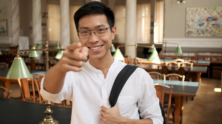 Junger, lächelnder asiatischer Student zeigt mit dem Zeigefinger in der Bibliothek