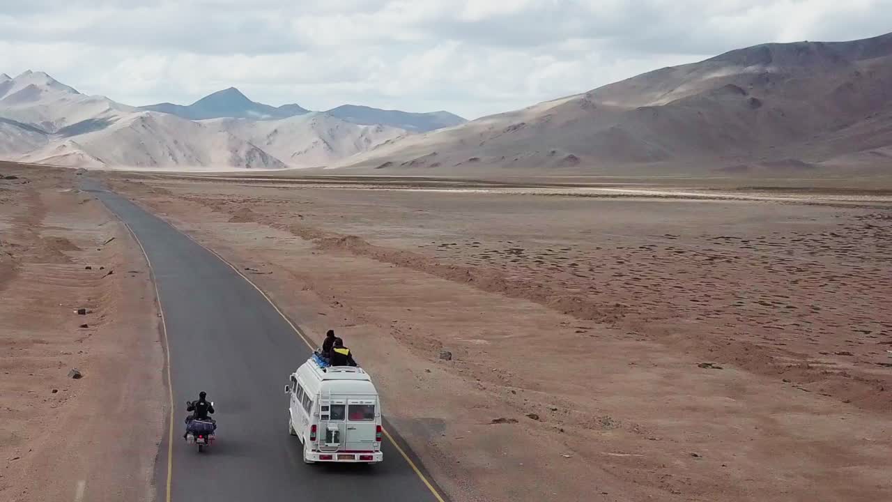 A lone cyclist stands on his bike and removes his helmet then is overtaken by two vans with people standing up and looking out of the vehicle roof.
