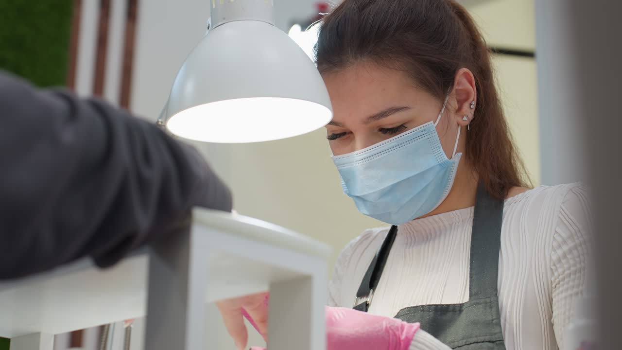 Nail technician with blond hair wearing pink gloves and blue mask carefully performing manicure on customer under white desk lamp in clean salon environment, demonstrating precision and concentration
