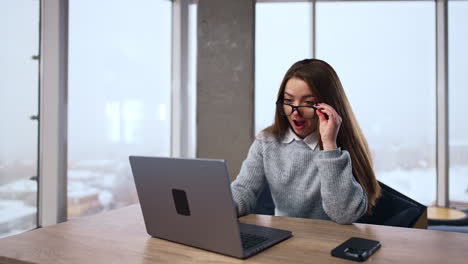 pretty brunette wearing glasses works on her laptop. woman looks at computer screen with surprise and puts off her glasses.
