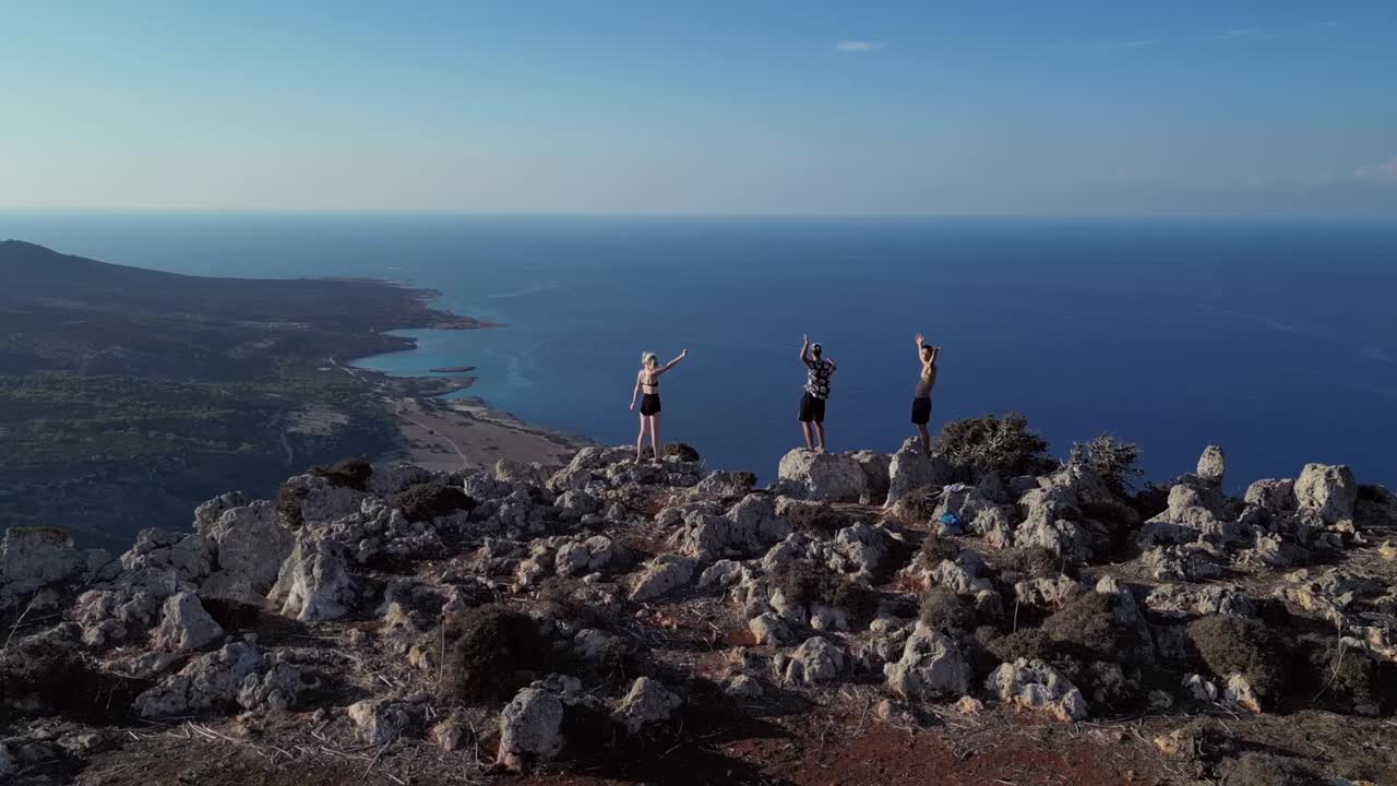 Drone captures two men and a woman standing on rugged rocks, overlooking the foggy Mediterranean Sea, surrounded by a misty, atmospheric seascape.