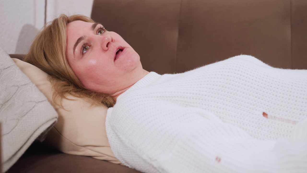 Guest lies on sofa facing professional and speaking softly with reflective gaze, pillow supports head, table beside holds clock, tissue box, pencils in warm calm therapy setting in private space