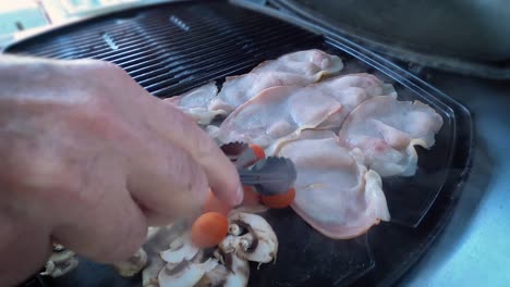 primer plano de una mano, girando tocino frito, champiñones y tomates con pinzas en una barbacoa moderna incorporada