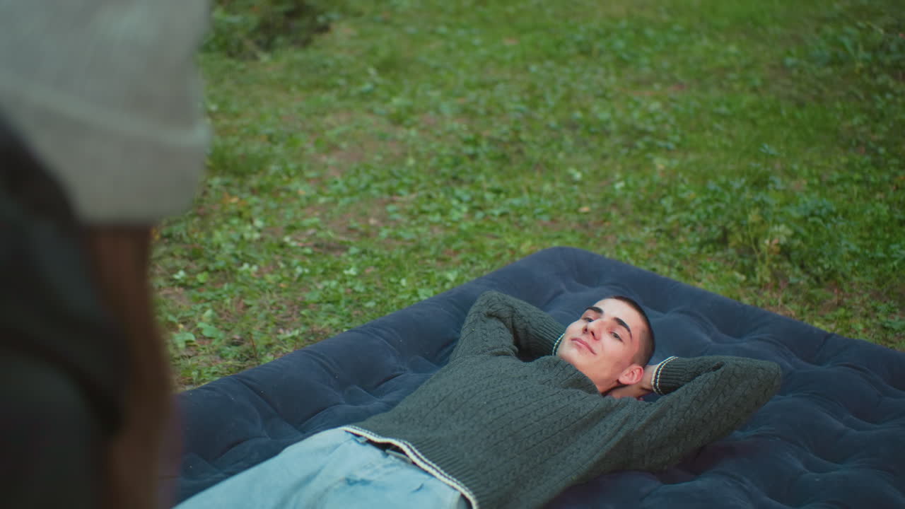 close up back view of woman bent over working while man relaxes on air bed with hands behind head smiling warmly at her in lush green forest campsite