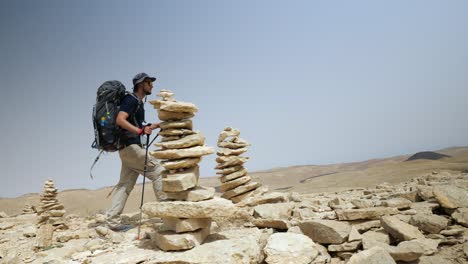 Vista-Estática-De-Un-Joven-Mochilero-Feliz-Caminando-En-El-Pico-De-La-Montaña-Entre-Piedras-Paradas-Una-Encima-De-La-Otra