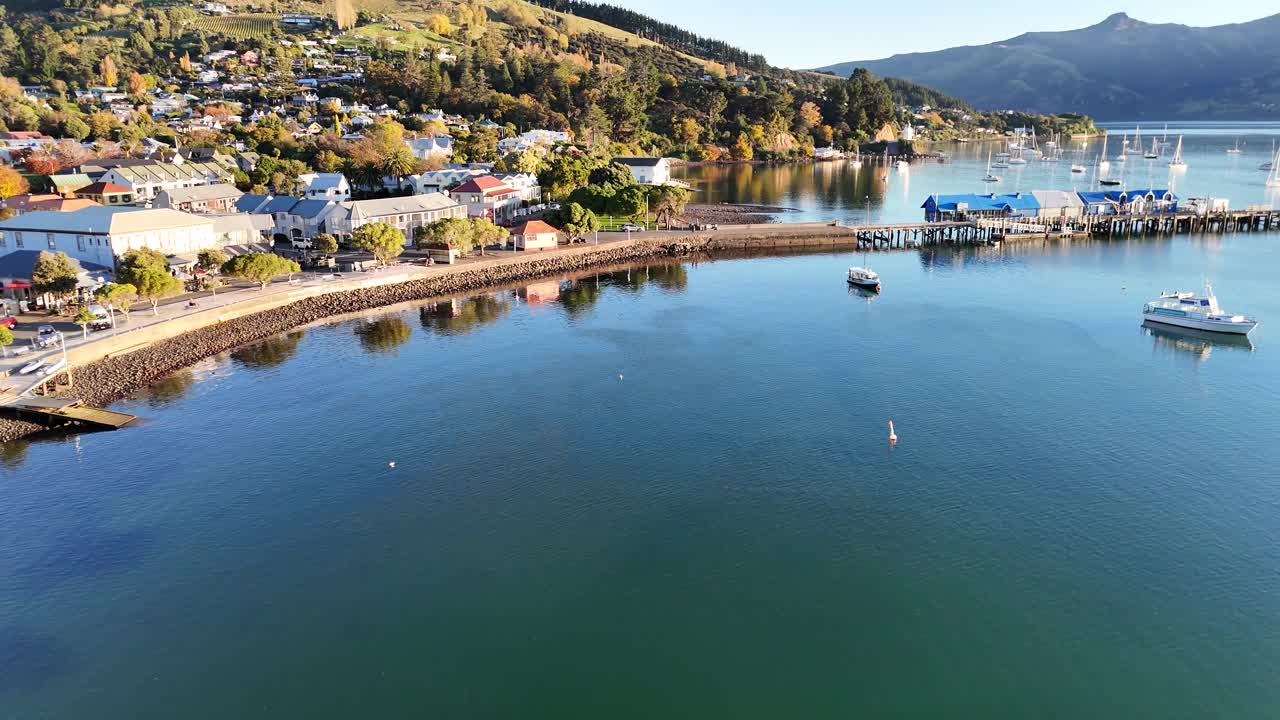 Drone footage captures Akaroa's serene harbor, showcasing boats, a pier, and surrounding hills under warm sunset lighting