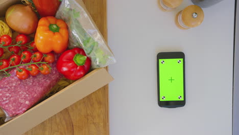 Toma-Cenital-De-Una-Caja-De-Ingredientes-Frescos-Junto-A-Un-Teléfono-Móvil-Con-Pantalla-Verde,-Filmada-En-Cámara-Lenta