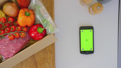 fotografía aérea de una caja de ingredientes frescos junto a un teléfono móvil de pantalla verde - tomada en cámara lenta