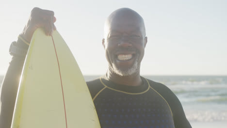 Portrait-of-happy-senior-african-american-man-holding-surfboard-at-beach,-in-slow-motion