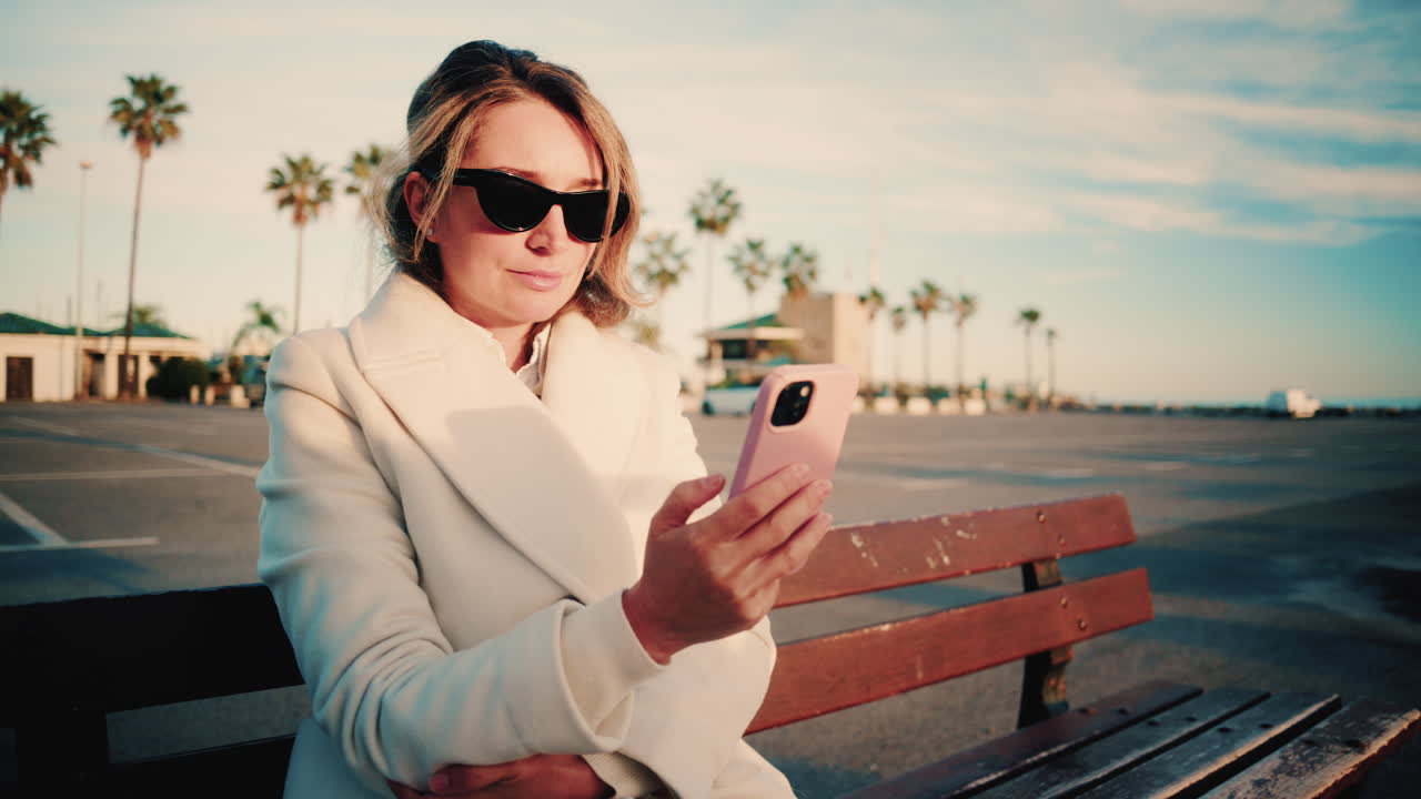 Donna elegante con cappotto bianco guarda il suo smartphone mentre è seduta su una panchina di legno in un parcheggio soleggiato costeggiato da palme