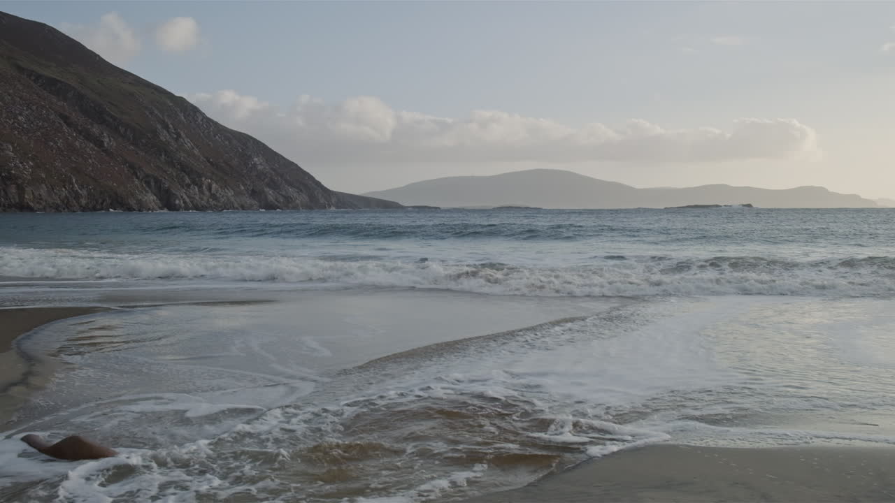 Panorama shot of a beach with water flowing back into the ocean after a wave. More waves hitting the beach. Clouds and mountain in the background.