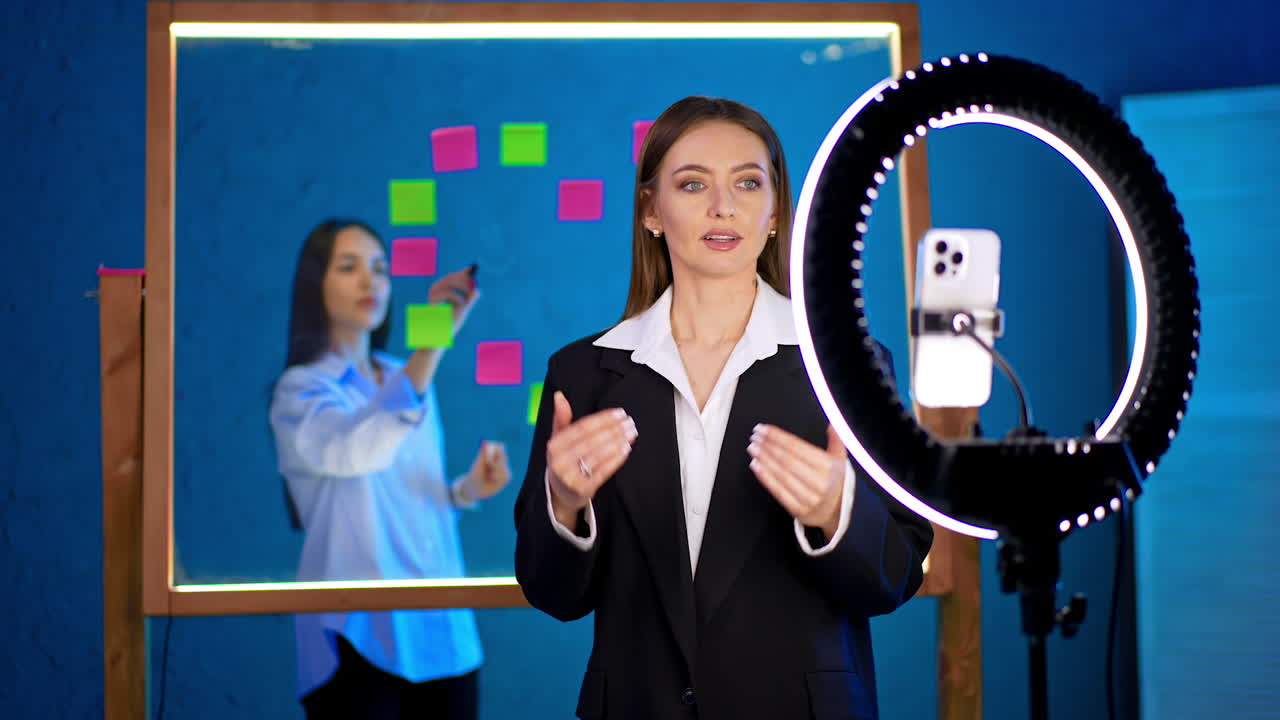 Woman presents in front of a ring light. A woman talks confidently to a camera while another person organizes sticky notes on a glass board in the background