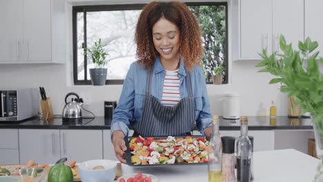 portrait of happy biracial woman in apron holding seasoned vegetables on baking tray, slow motion