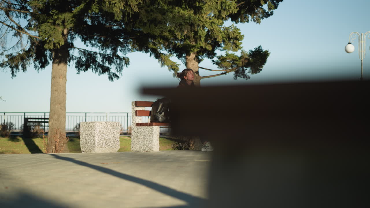 una niña se sienta sola en un banco en un parque urbano, mirando triste y perdida en sus pensamientos. lleva un abrigo marrón, pantalones vaqueros azules y zapatos blancos, con una mochila negra colocada a su lado