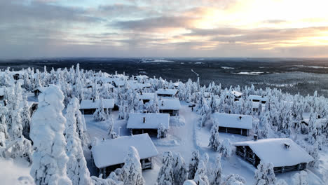 flying over snowy trees, toward a cottage village on top a fell, winter sunset in lapland - aerial view