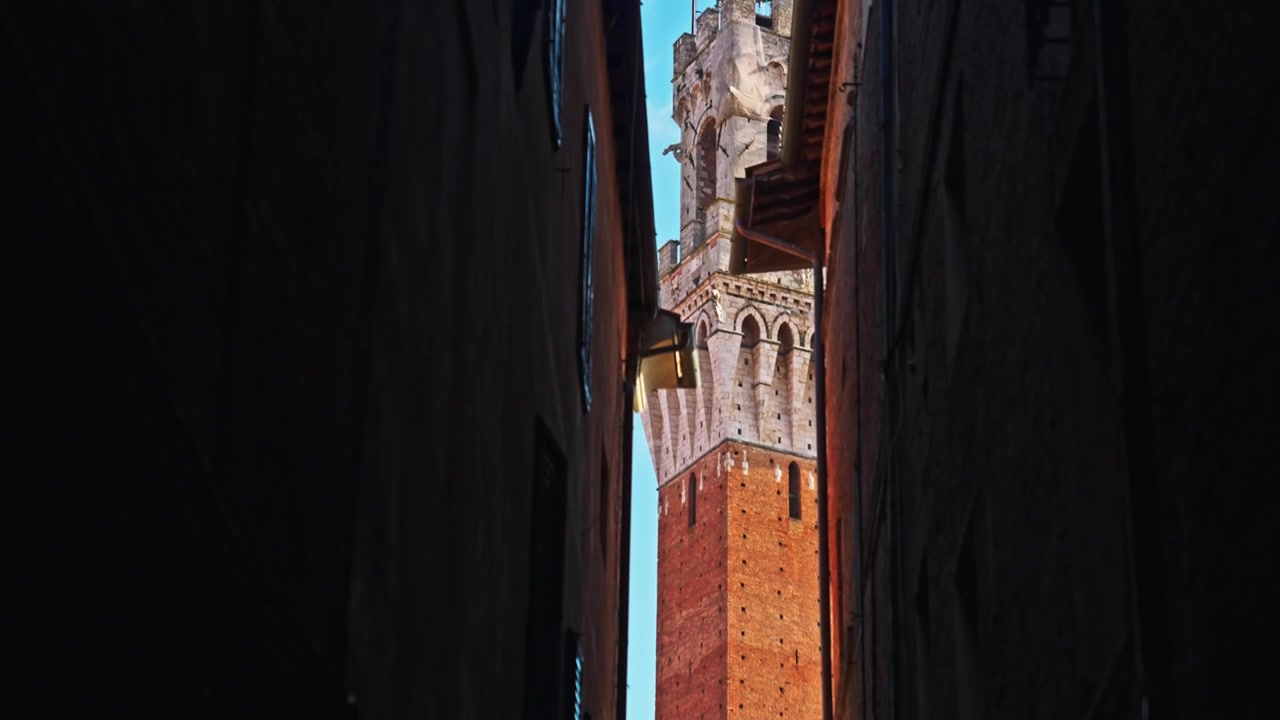 La Torre del Mangia si intravede tra uno stretto varco negli edifici vicino a Piazza del Campo, Siena, Italia. Ripresa in elevazione a mano