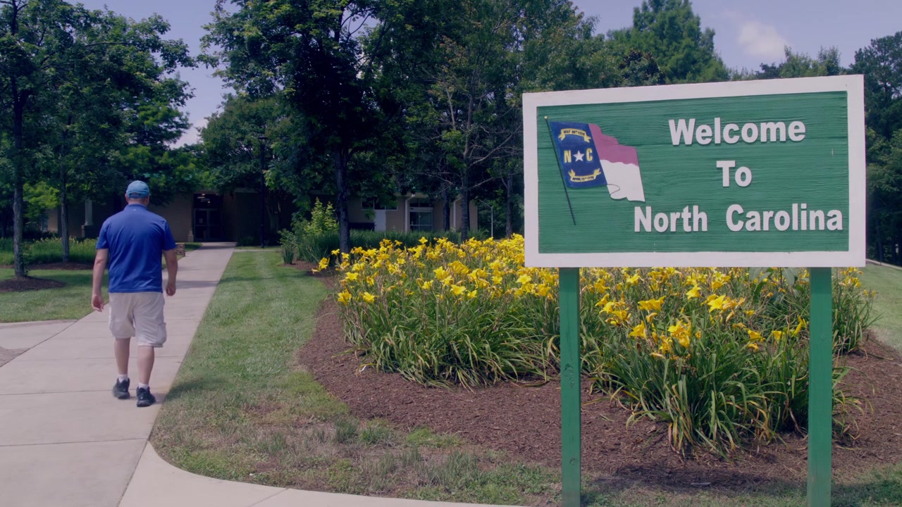 Uomo che cammina accanto al cartello "Benvenuti in Carolina del Nord" in un'area di sosta, con video stabile.