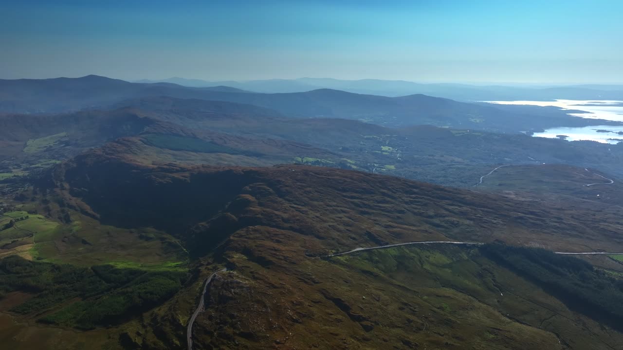 Caha Pass, County Cork, Ireland, September 2024. Drone tracks right in a panoramic high angle overview of a winding road crossing the rugged mountain ridge and descending to the North Atlantic Ocean.