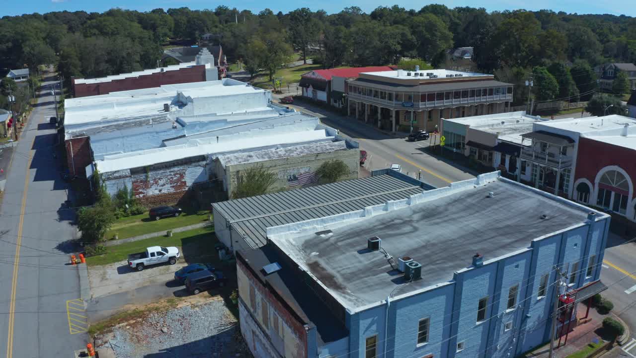 A Quiet Small Town Street is Lined With Old-style Buildings and Surrounded by Greenery Under a Clear Blue Sky - Aerial Drone Shot