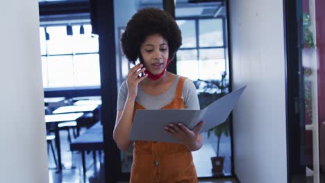 Mixed-race-businesswoman-wearing-mask-walking-going-through-paperwork-in-modern-office