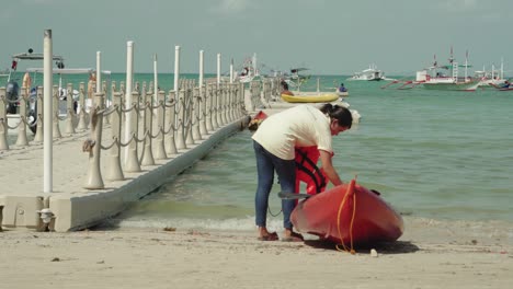a female beach worker taking out two red life vests and paddles from a kayak for inventory and rental in el nido, philippines - wide shot