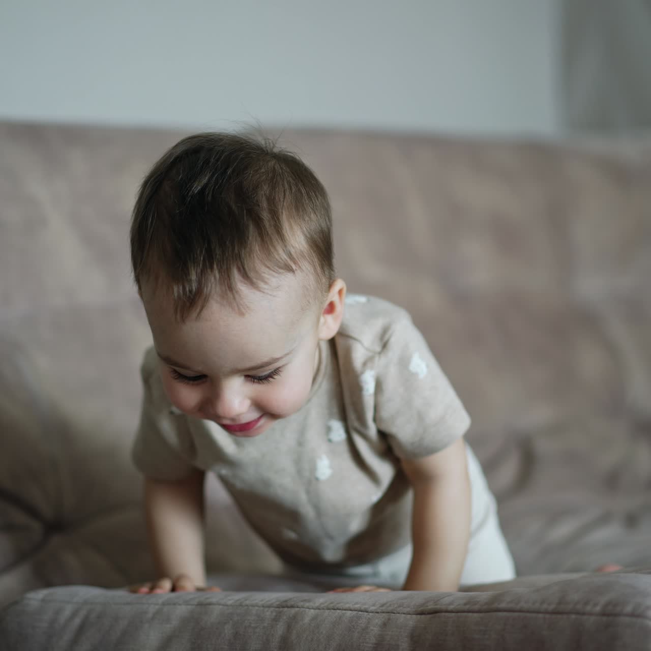 Happy active toddler playing hide and seek on the sofa. Kid lies down and rises above the pillow and laughs adorably