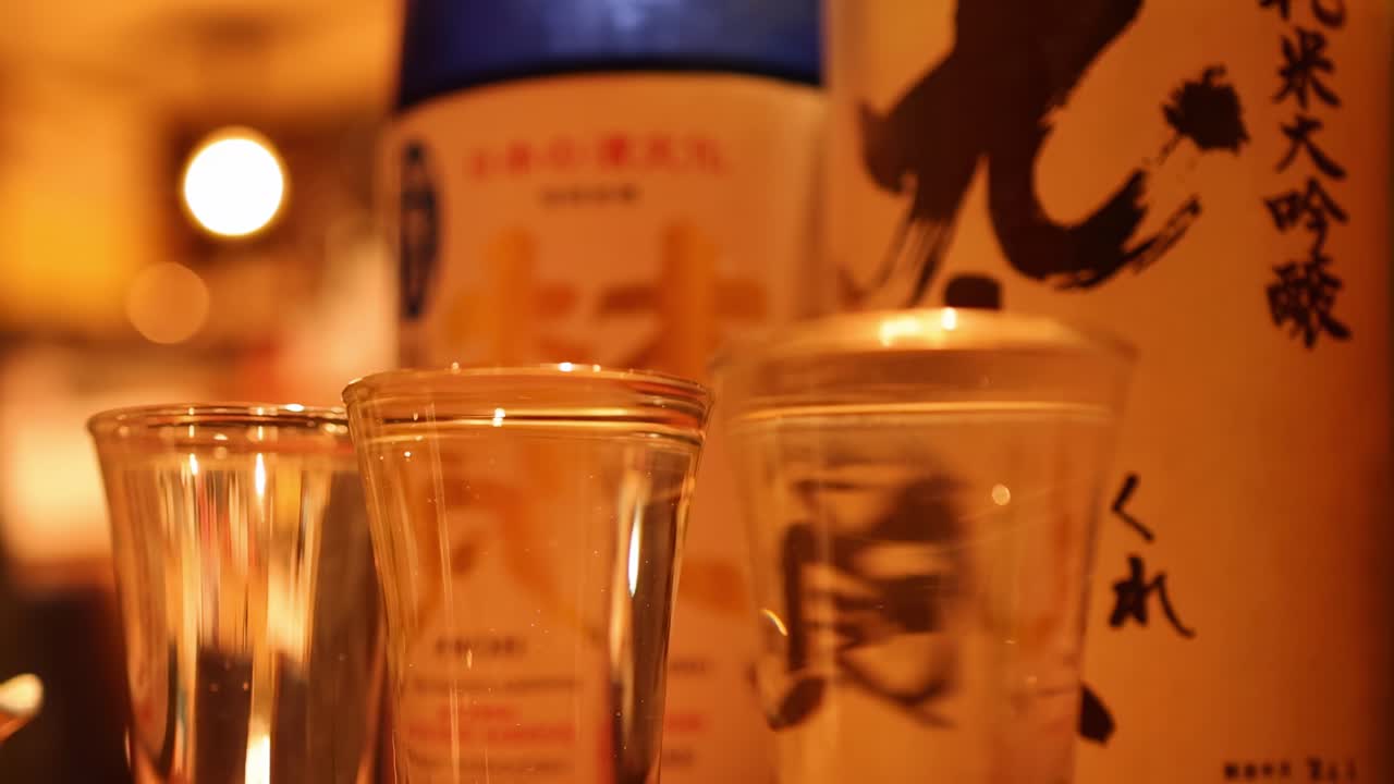 Detailed view of sake bottles and glasses with warm lighting in a bar setting.