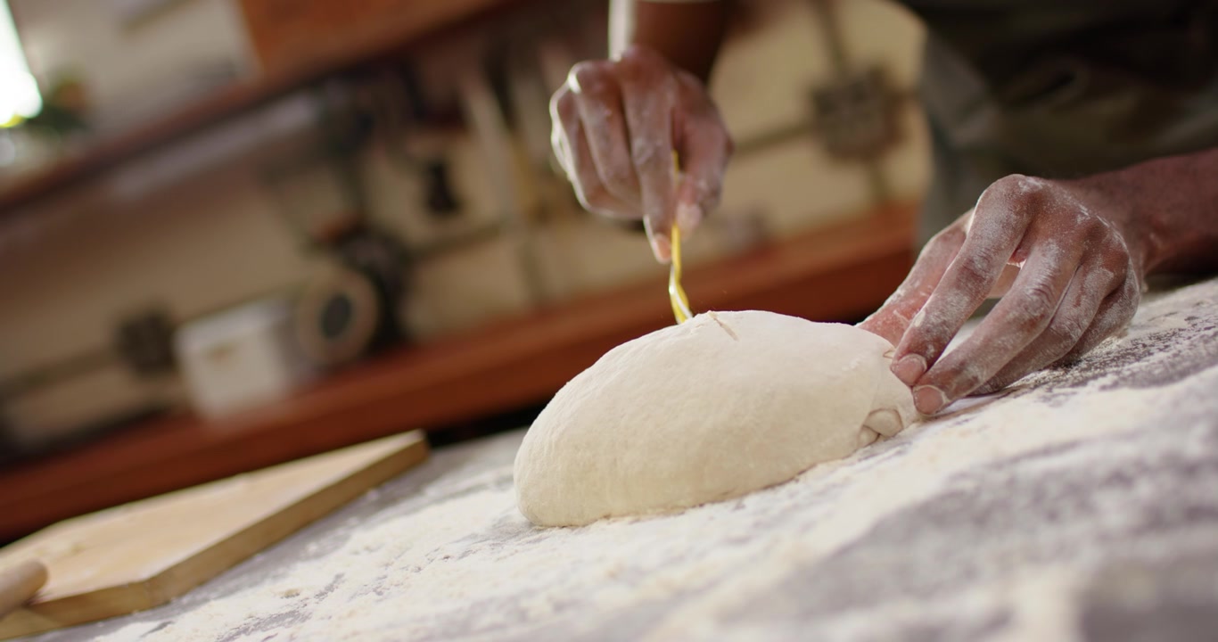 Male baker noticing proofed dough, scoring loaf with yellow tool on floured wood counter for baking