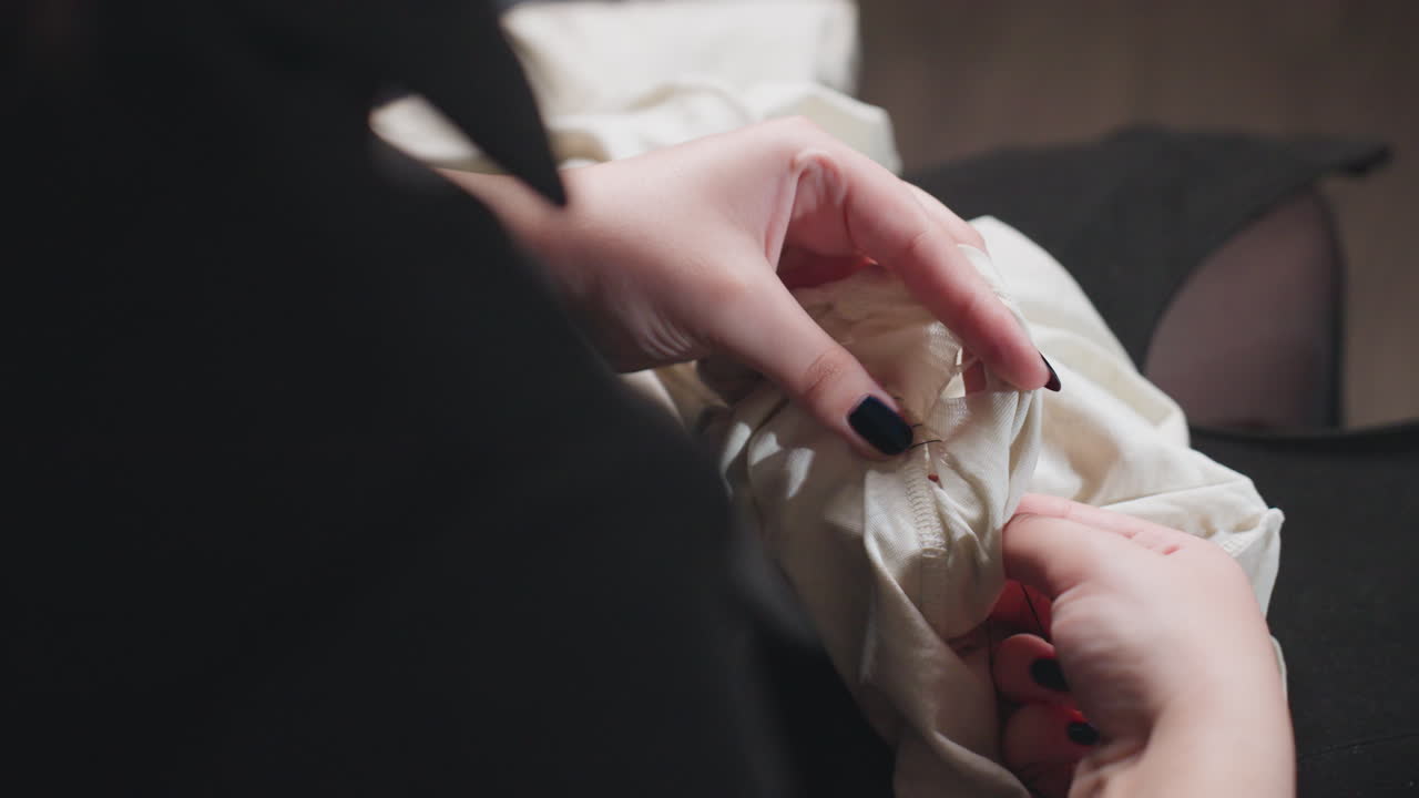 Female hands stitch torn white dress with needle and thread, steady work on lap, polished nails, black outfit, indoor focus, careful repair of fabric seam in soft home light