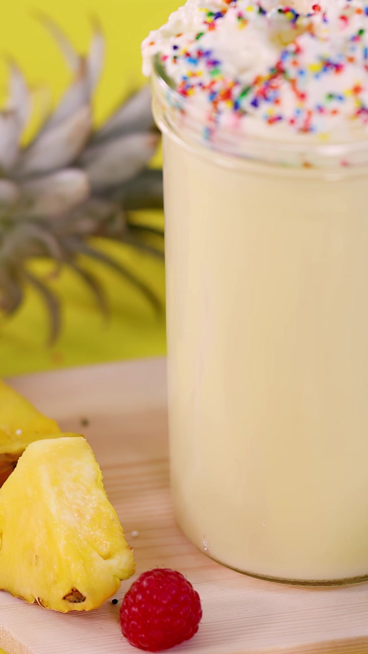 Bicchiere di frullato all'ananas che ruota con panna montata, guarnizione di frutta fresca, sfondo da studio giallo brillante