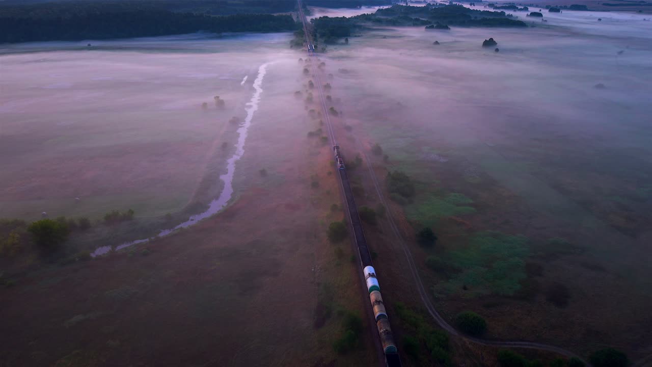 Aerial view, freight train moving along railway tracks covered in morning fog near the Nida River, Poland, with peaceful meadows and countryside landscape
