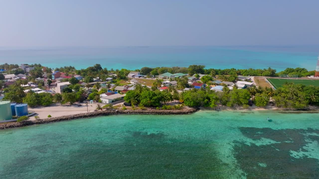 Luftoptagelser fra drone, der flyver parallelt med Vashafaru-øens sydlige kystlinje, med det lokale fodboldfelt, havneområdet og turkise tropiske farvande i det nordlige Maldiverne.