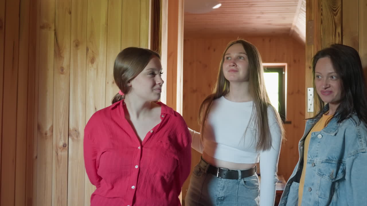 three students step into wooden lounge with natural lighting and cozy atmosphere, one in red shirt examining wall while others observe surroundings