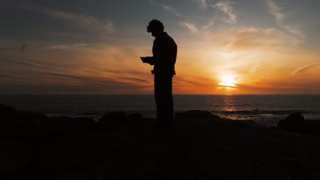 silhouette of man standing and reading book at sunset by the ocean. muxia, spain. orbiting shot