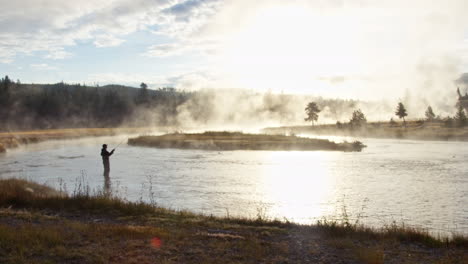 Side-angle-silhouette-of-a-man-with-fishing-rod-in-his-hand-standing-in-river-illuminated-by-shiny-sunlight-and-smoke,-POV