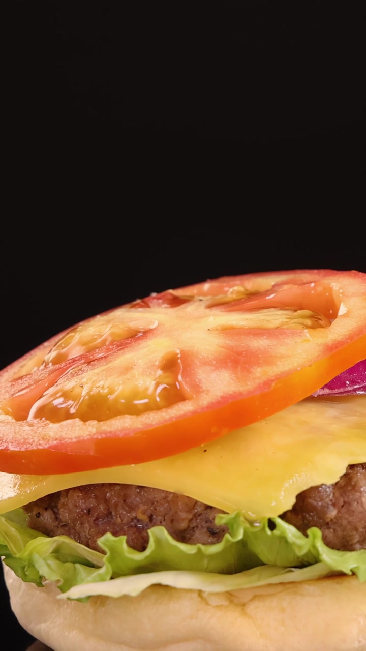 Un panino con semi di sesamo cade al rallentatore su un cheeseburger di manzo con lattuga, pomodoro, cipolla e cheddar, su uno sfondo nero pulito con illuminazione da studio.