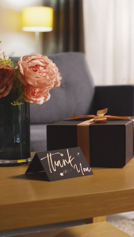 vertical video of present in gift wrapped box decorated with ribbon with thank you card and flowers on table in lounge