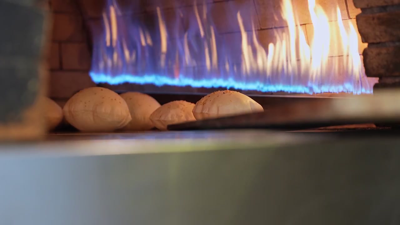 Primo piano cinematografico di pane pita fresco che cuoce in un forno a pietra tradizionale con fiamme blu. Una pala di legno entra nell'inquadratura, perfetto per il marketing di ristoranti e lo storytelling culinario.