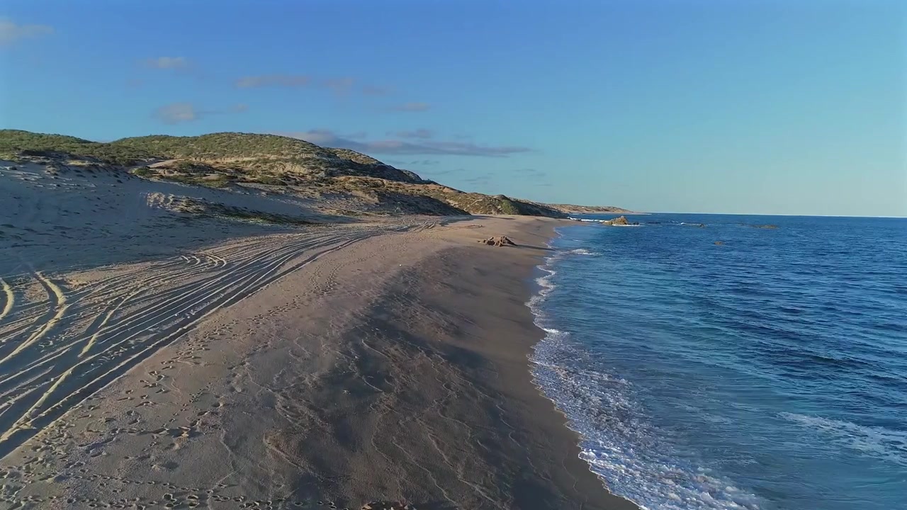Drone flyver lavt over stranden ved Playa El Rinconcito på Østkappen af Baja California Sur, observerer bølger rulle ind på kysten med klitter til venstre og havet til højre, mens den holder en lav højde